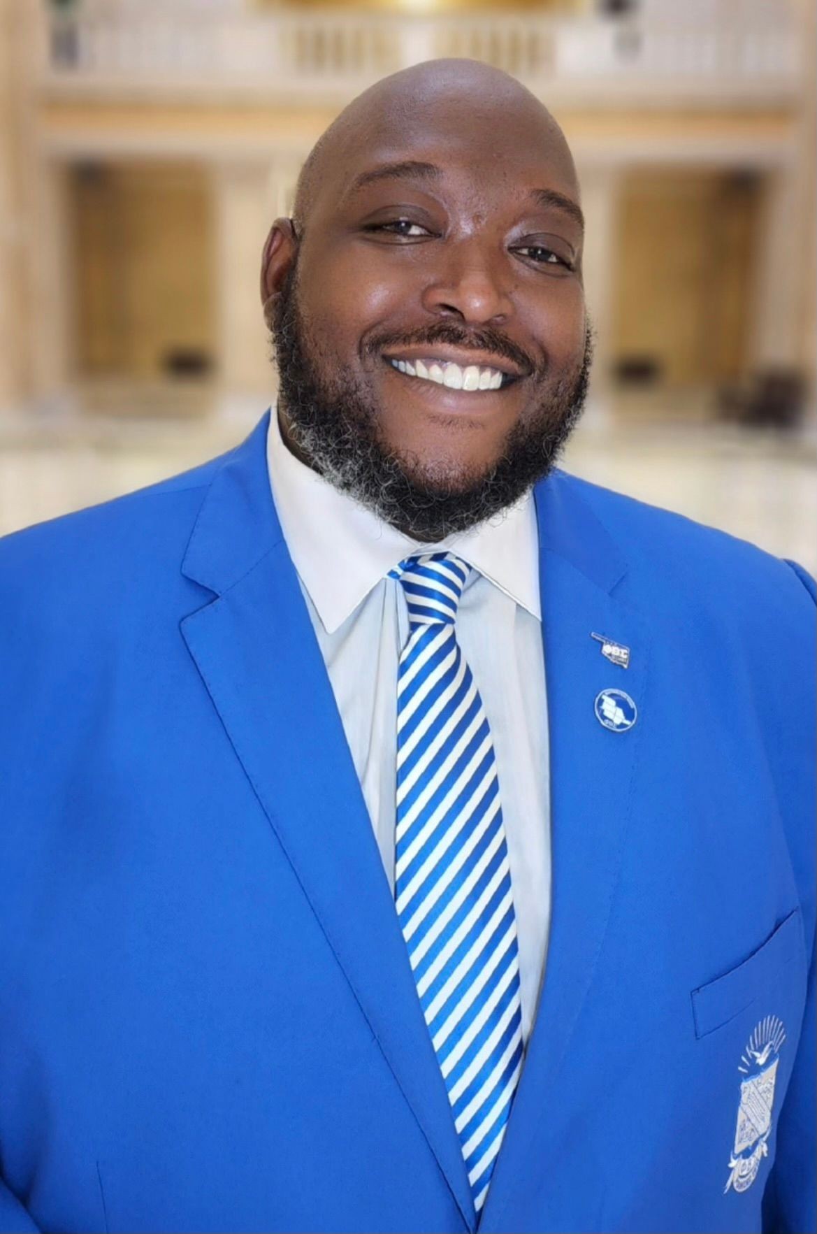 Smiling man in a blue suit with matching striped tie, standing in a blurred indoor setting.